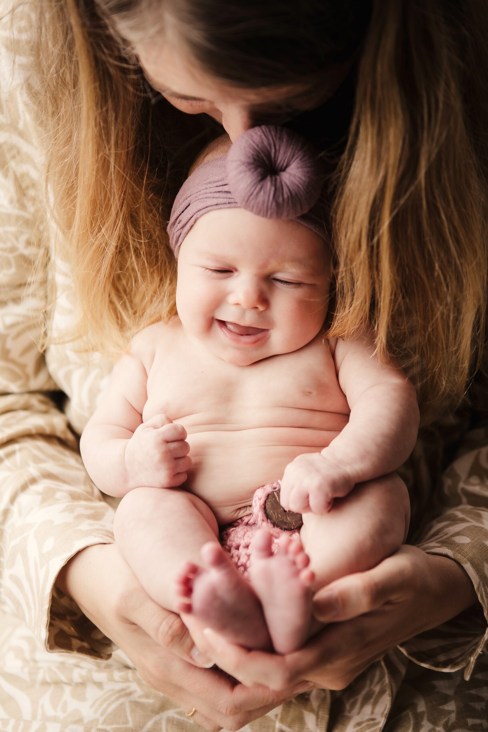 St. Louis newborn photographer, mother kissing the head of her laughing baby girl in a mauve bow headband, tiny feet cradled in her hands