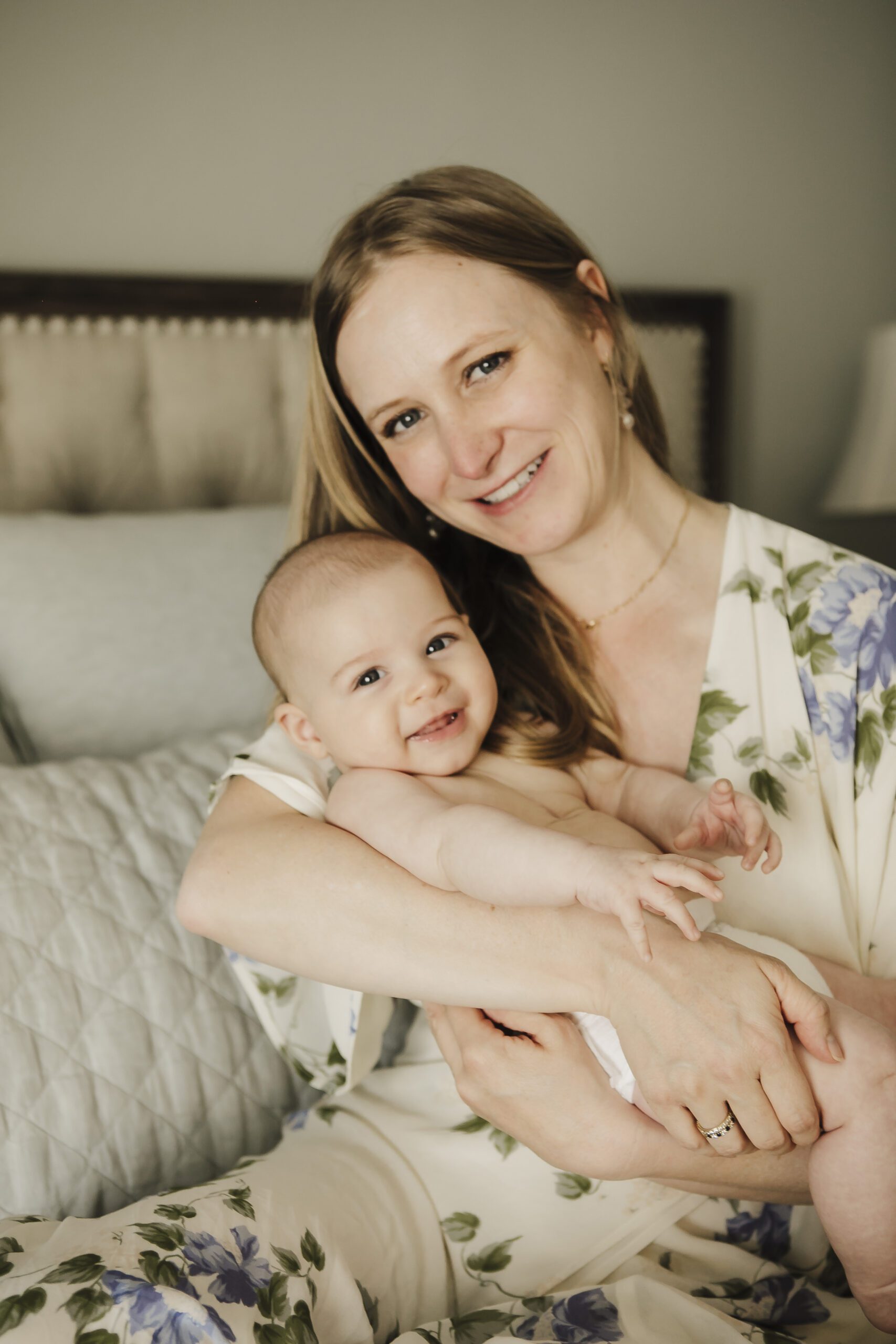 St. Louis newborn photographer, blonde mother in a blue floral dress smiling with her baby boy held against her chest on a bed
