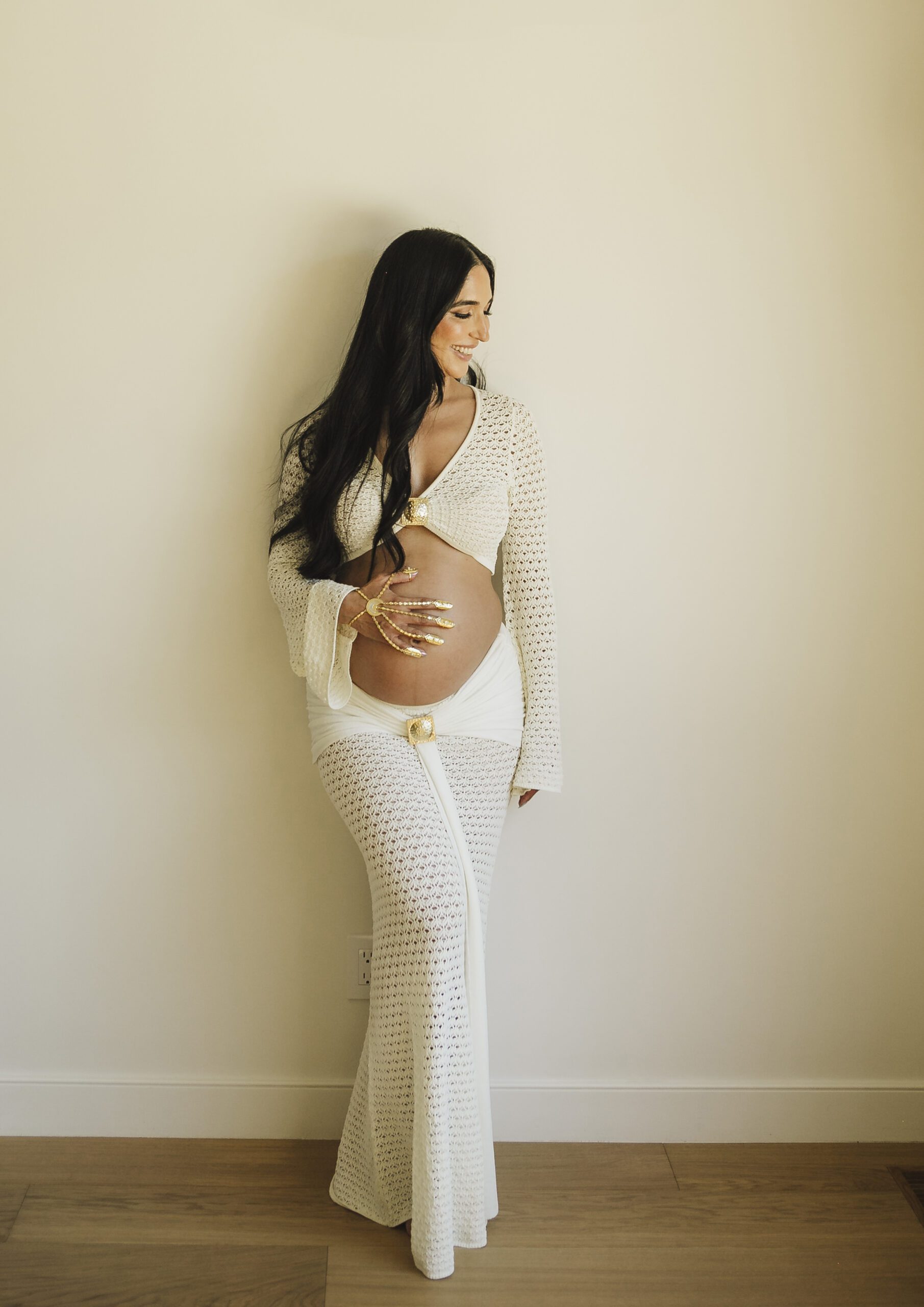 St. Louis maternity photographer, expecting mother in a cream crochet set with ornate gold hand jewelry over the bare bump against a plain studio wall