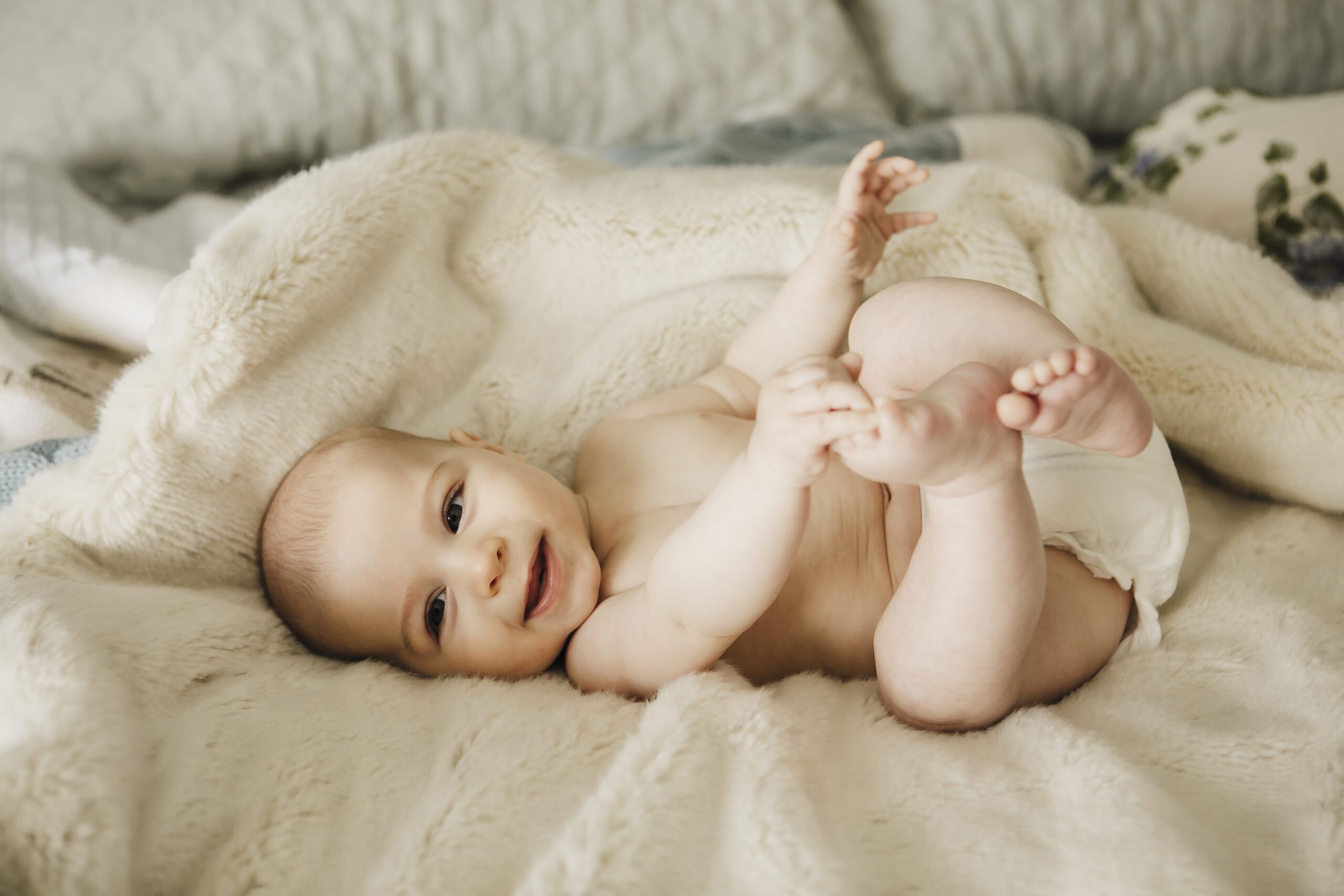 Baby smiling and holding its feet while lying on a soft cream blanket in a bright room