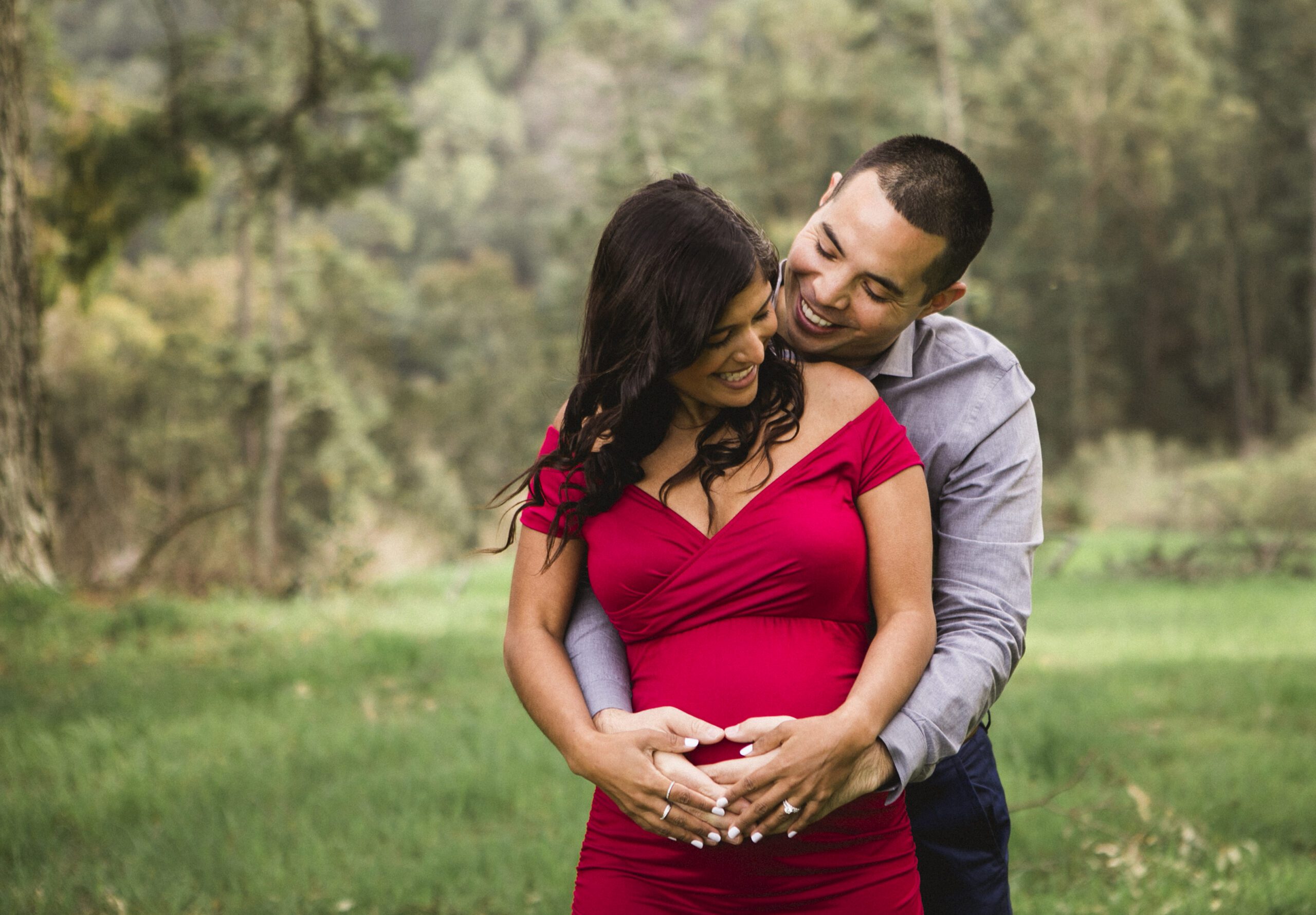 St. Louis maternity photographer, expecting couple embracing in a grassy meadow, mother to be in a red gown with hands cradling the belly