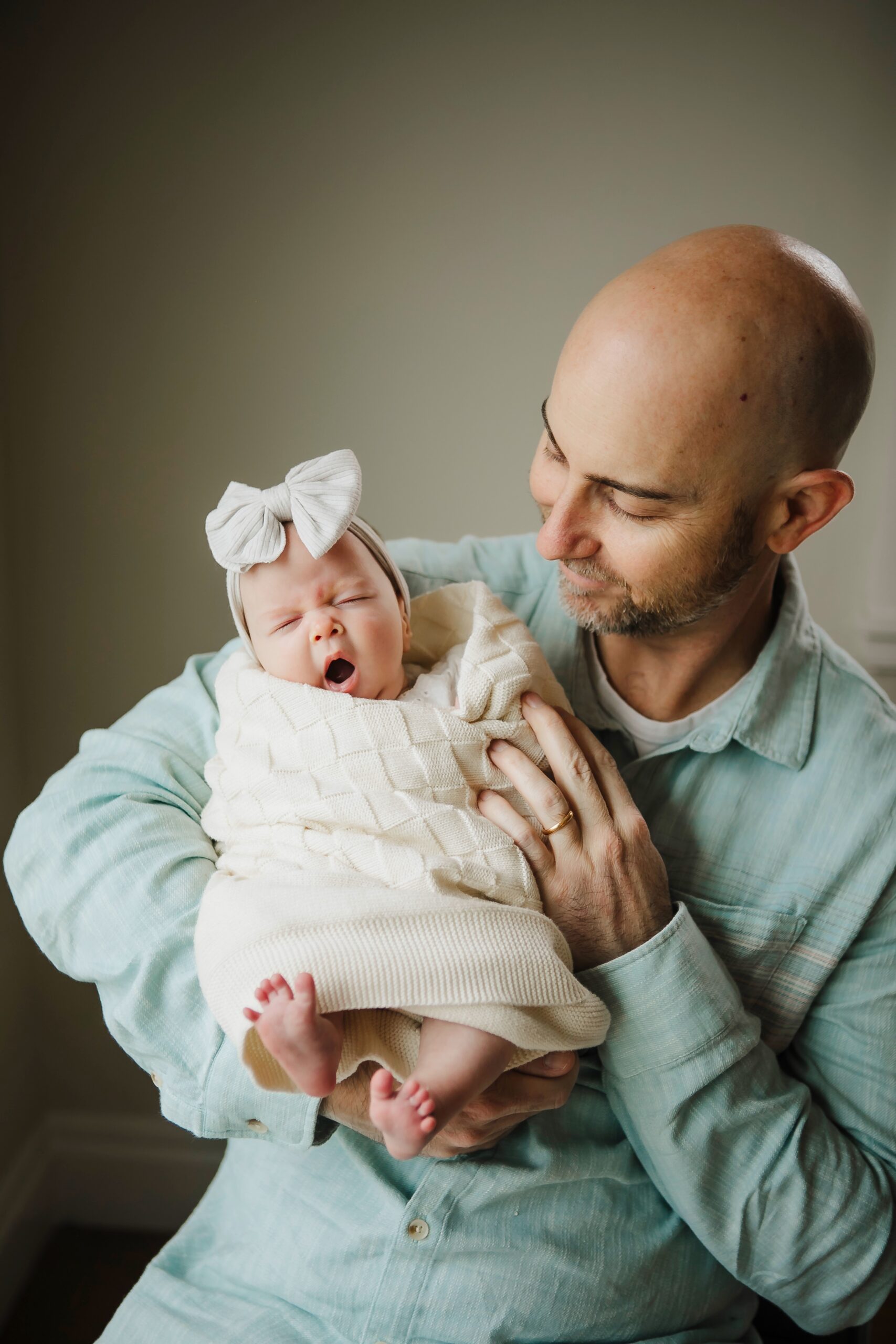 St. Louis newborn photographer, father in a sage shirt holding his yawning baby girl wrapped in a cream cable knit with a white bow headband