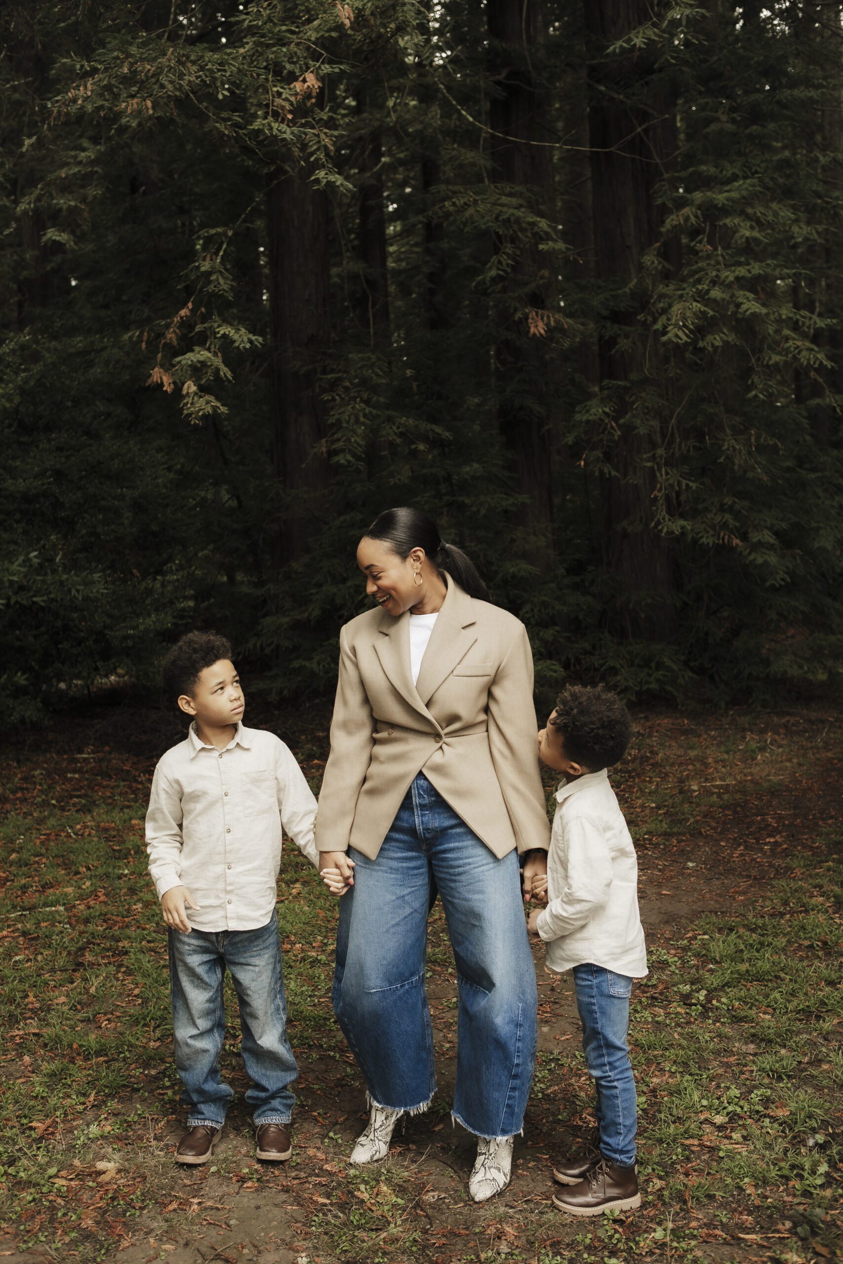 St. Louis family photographer, mother in a tan blazer holding hands with her two young sons in a forest clearing