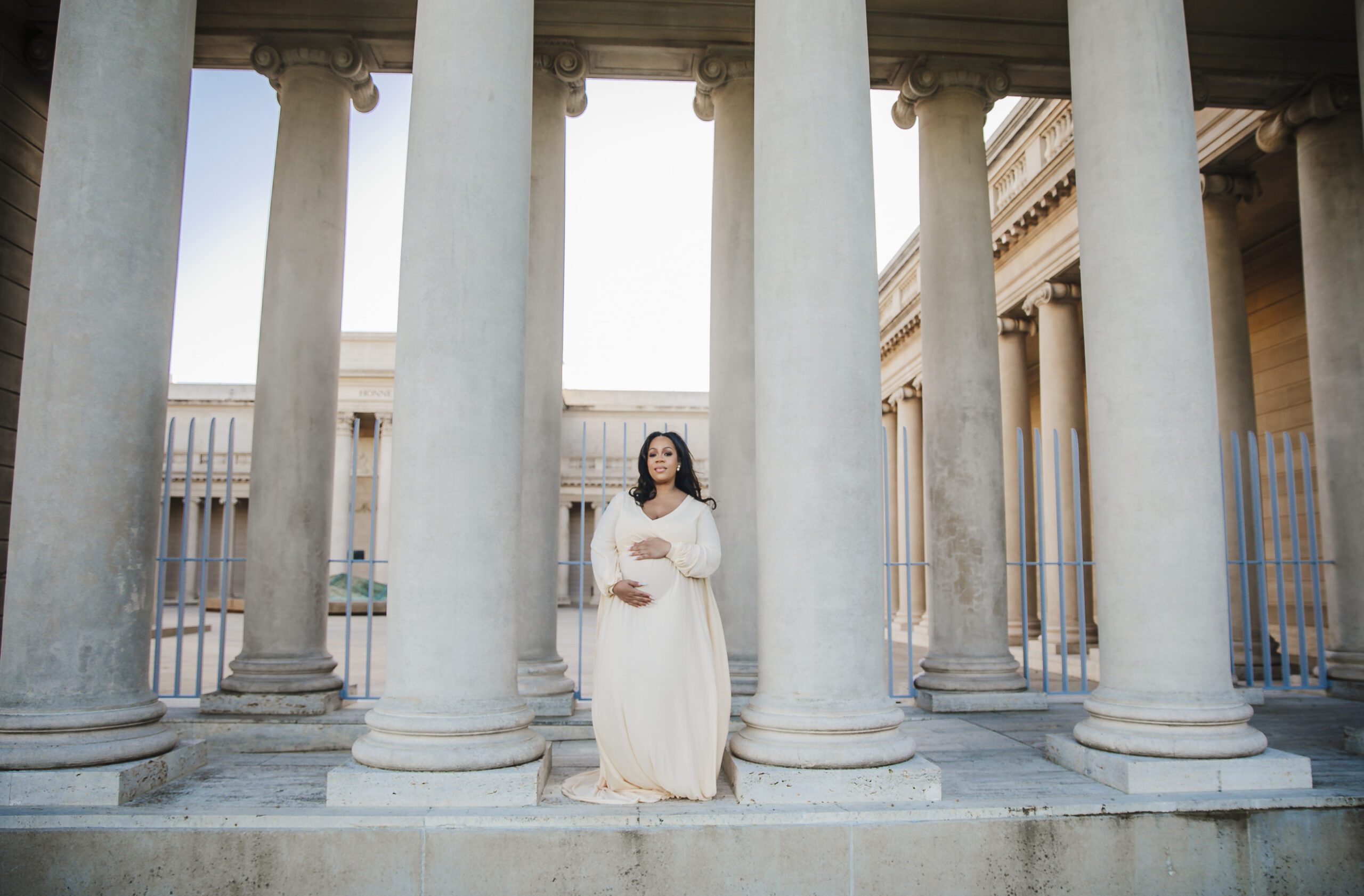 Maternity portrait of a pregnant woman in a flowing cream gown standing between tall stone columns of a neoclassical colonnade