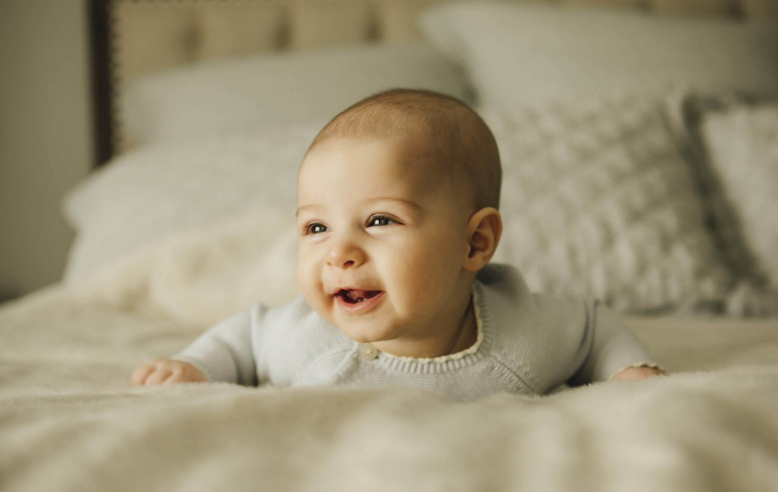 Smiling baby in a pale blue knit outfit doing tummy time on a bed with soft neutral bedding