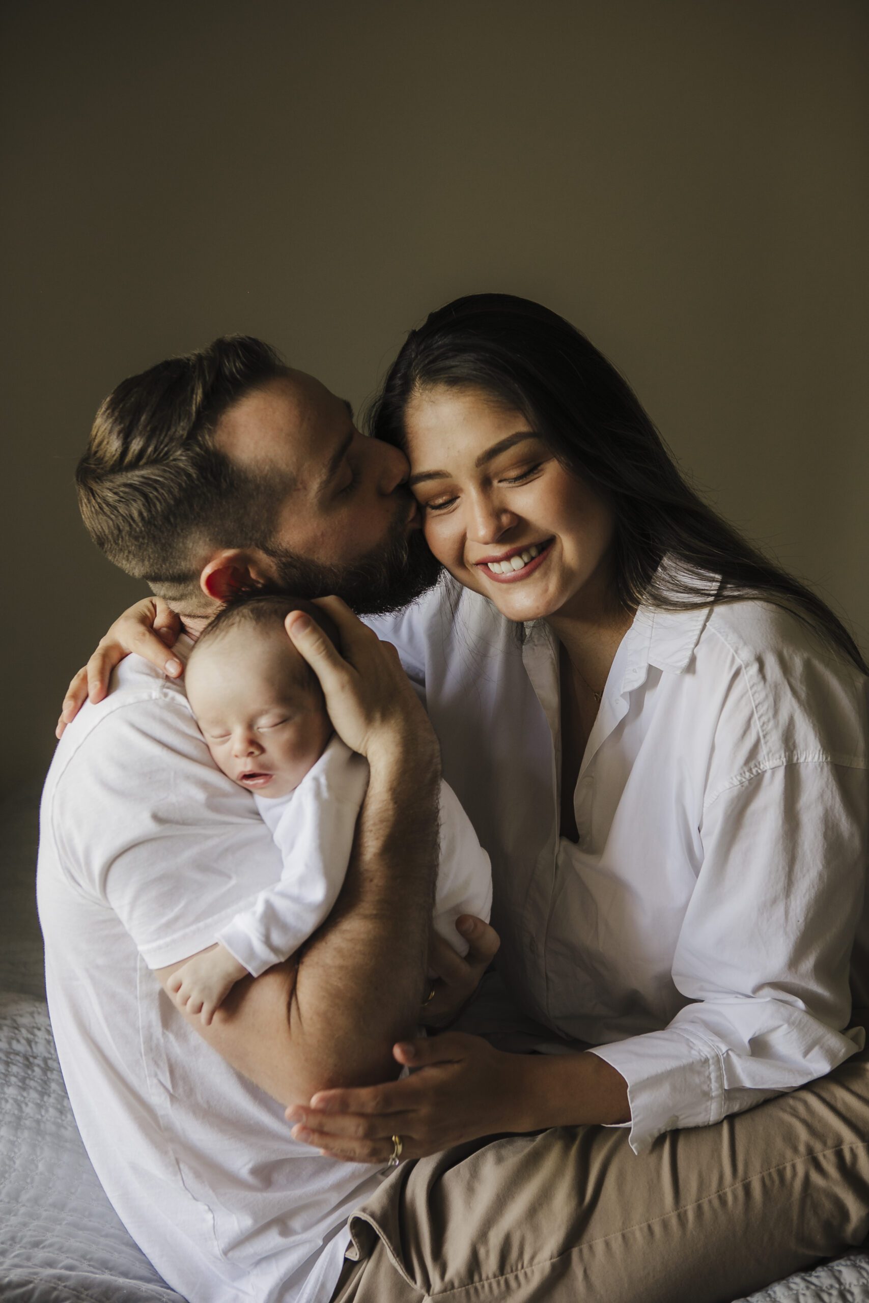 St. Louis newborn photographer, new mother smiling as the father kisses her temple and cradles their sleeping newborn dressed in white