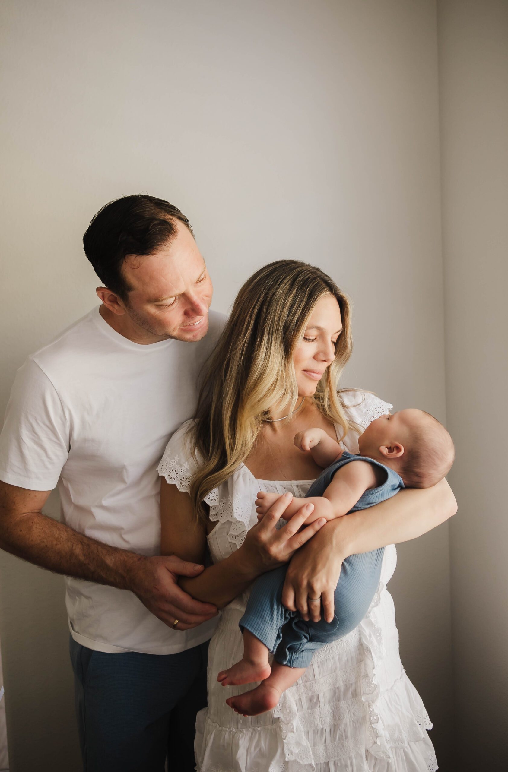 St. Louis newborn photographer, parents dressed in white holding their newborn in a blue outfit by a window