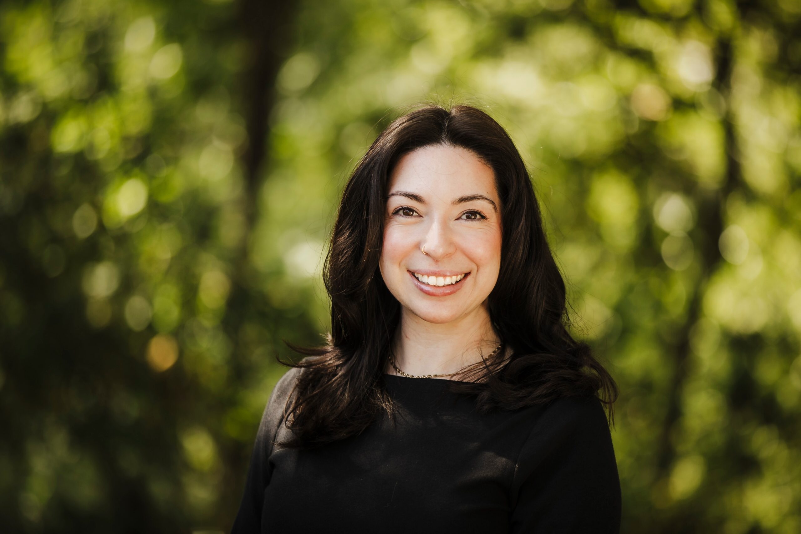 Professional headshot by a St. Louis headshot photographer, smiling woman with long dark wavy hair in a black top against a green leafy background