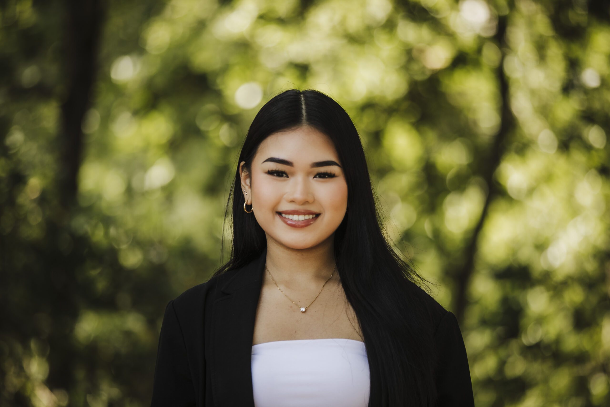 St. Louis headshot photographer, smiling young woman with long dark hair in a black blazer over a white top, outdoors against soft green foliage