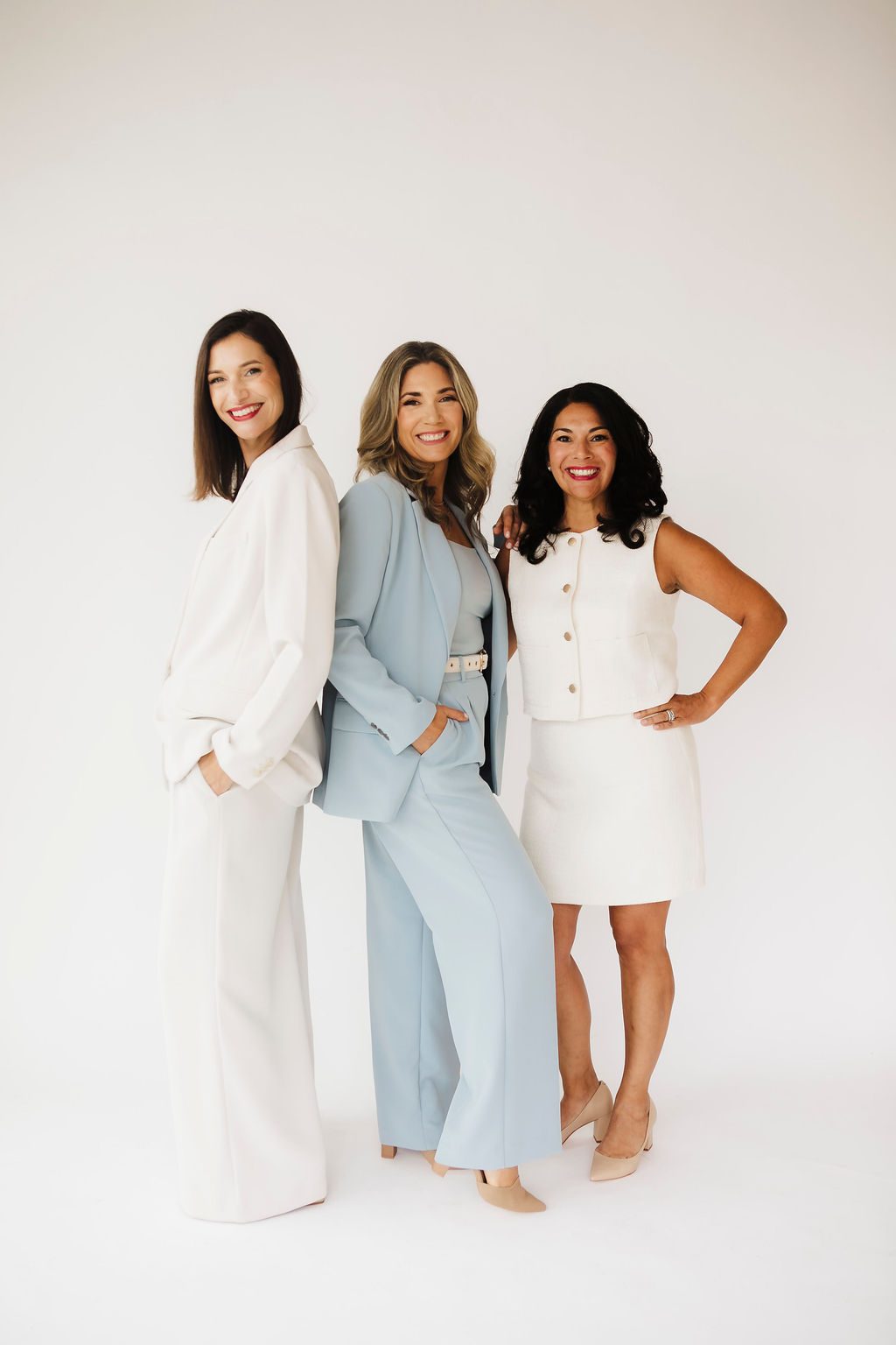 Team branding portrait of three smiling women in tailored suits, one in cream, one in light blue and one in white, standing together against a light studio backdrop