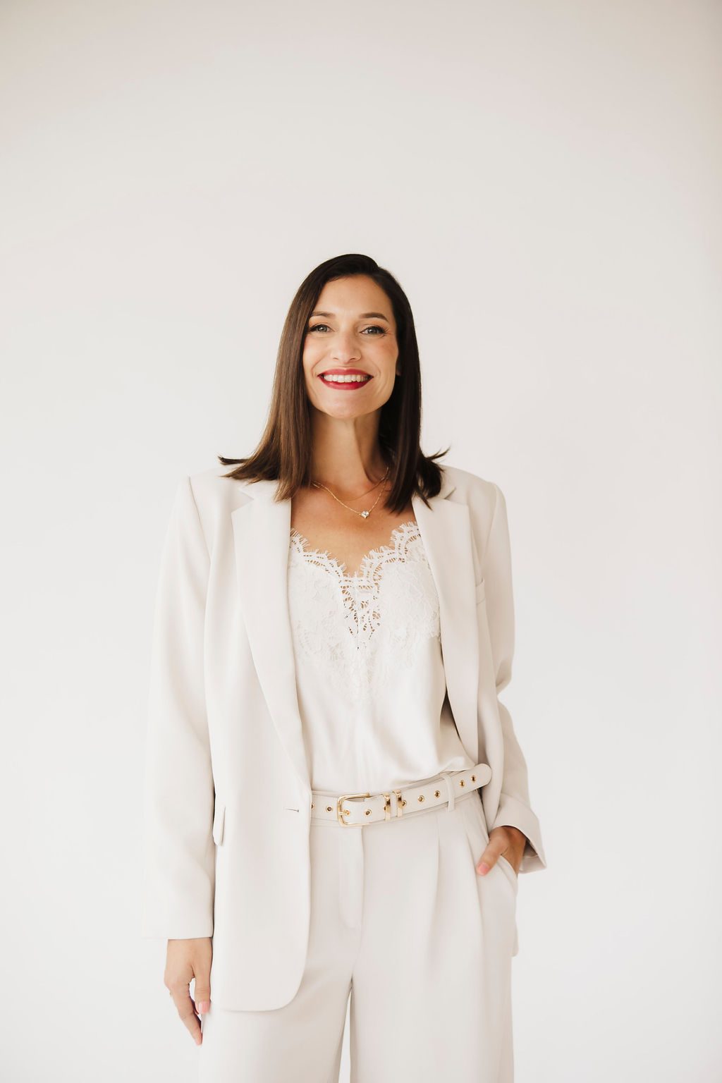 Personal branding portrait of a smiling woman with dark hair in a cream blazer, white lace camisole and belted trousers, hands in her pockets, against a light studio backdrop