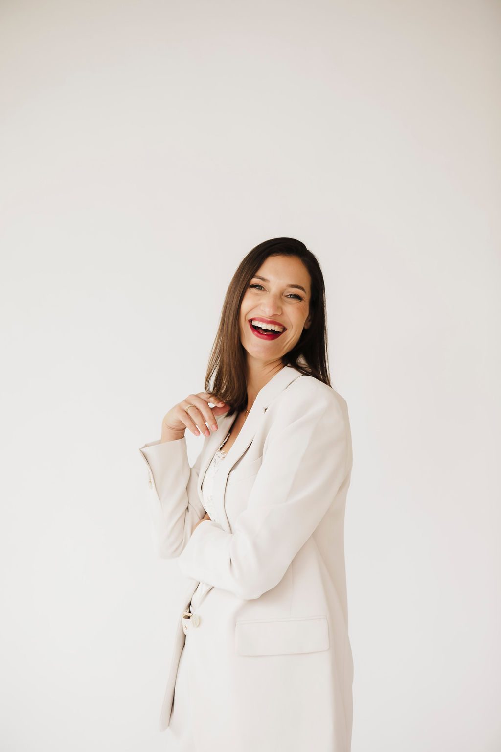 Personal branding portrait of a laughing woman with long dark hair in a beige blazer over a white lace top, against a light studio backdrop