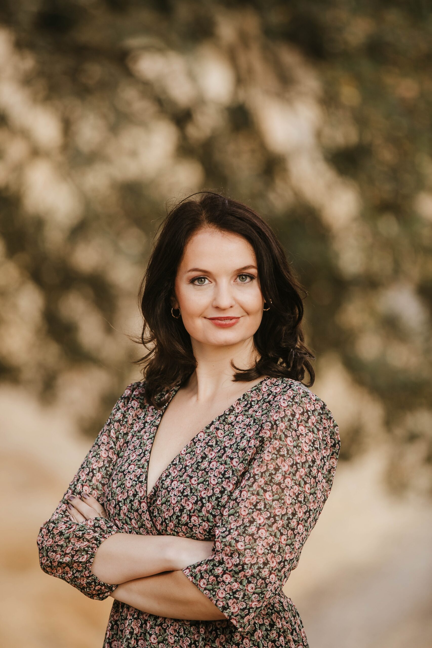 St. Louis headshot photographer, woman with dark hair in a floral dress standing with arms crossed against a soft, warm outdoor backdrop